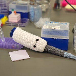 May include: A white crocheted pipette with a gray and blue tail, with black eyes and a pink mouth, lying on a gray surface. A clear plastic container with a blue lid and a clear glass jar with a white lid are in the background.