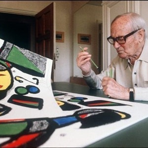 May include: An older man wearing glasses and a white shirt is working on a large, colorful abstract painting. The painting is on a table and the man is using a tool to add details to the artwork.