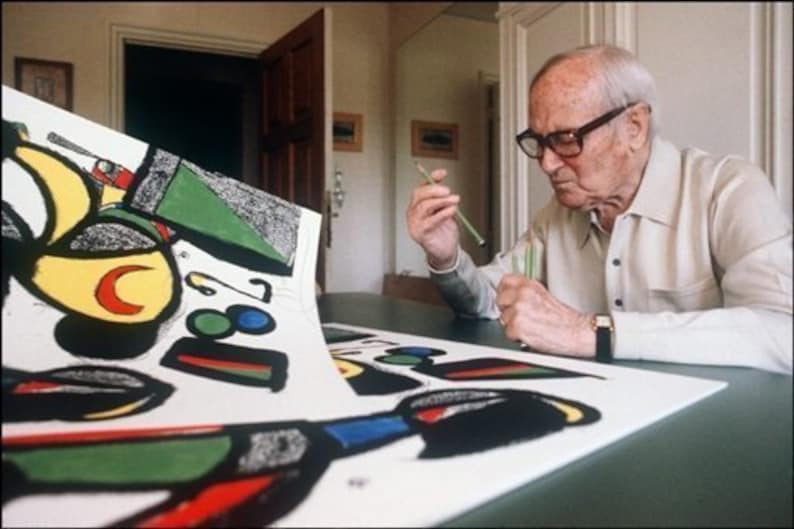 May include: An older man wearing glasses and a white shirt is working on a large, colorful abstract painting. The painting is on a table and the man is using a tool to add details to the artwork.