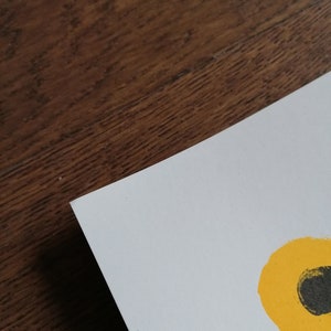 K&ouml;nnte beinhalten: Eine wei&szlig;e Papierecke mit einem gelb-schwarzen abstrakten Design. Das Papier liegt auf einer dunkelbraunen Holzoberfl&auml;che. Das Design zeigt einen gelben Kreis mit einem schwarzen Oval darin.