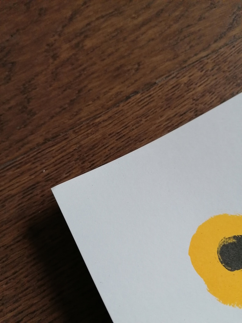 K&ouml;nnte beinhalten: Eine wei&szlig;e Papierecke mit einem gelb-schwarzen abstrakten Design. Das Papier liegt auf einer dunkelbraunen Holzoberfl&auml;che. Das Design zeigt einen gelben Kreis mit einem schwarzen Oval darin.