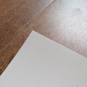 K&ouml;nnte beinhalten: Eine Ecke wei&szlig;en Papiers mit schwarzem Text auf einer Holzoberfl&auml;che. Die Kante des Papiers ist sauber und kontrastiert mit der Holzmaserung.