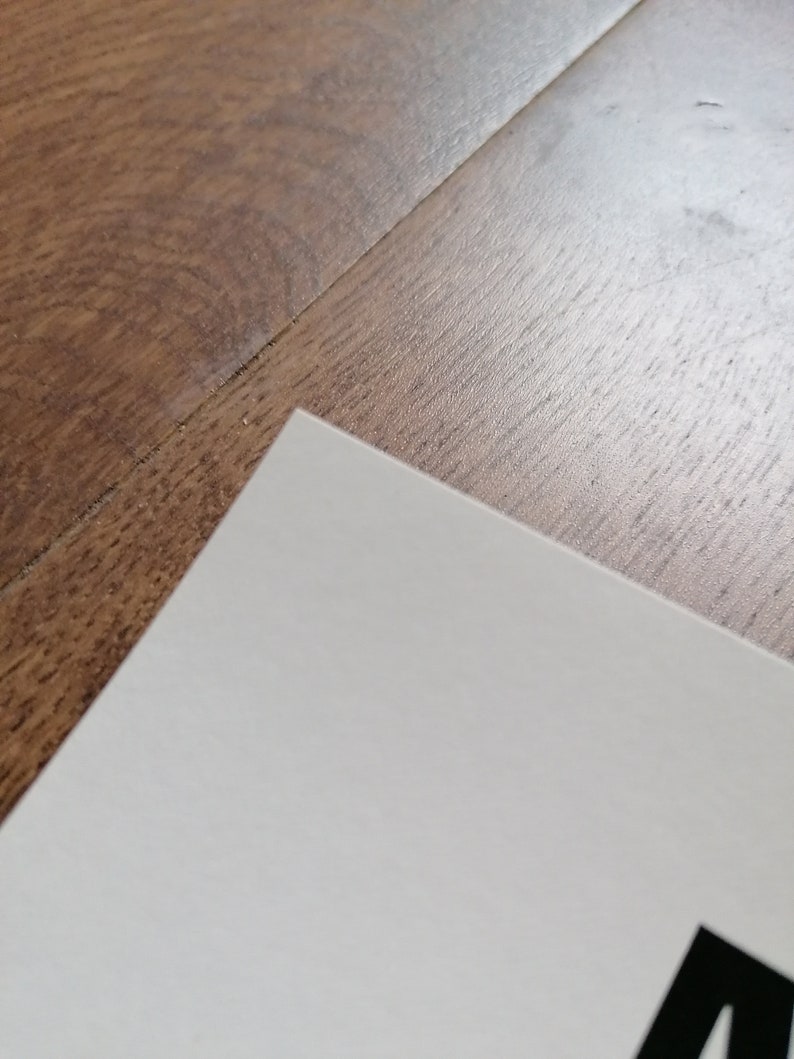 K&ouml;nnte beinhalten: Eine Ecke wei&szlig;en Papiers mit schwarzem Text auf einer Holzoberfl&auml;che. Die Kante des Papiers ist sauber und kontrastiert mit der Holzmaserung.