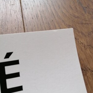 K&ouml;nnte beinhalten: Eine Ecke eines wei&szlig;en Papiers mit dem Buchstaben "&Eacute;" in Schwarz. Das Papier liegt auf einer Holzoberfl&auml;che.