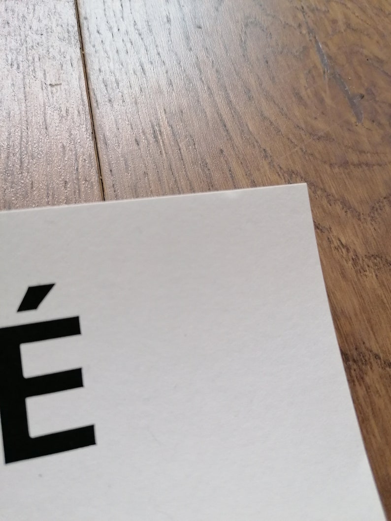 K&ouml;nnte beinhalten: Eine Ecke eines wei&szlig;en Papiers mit dem Buchstaben "&Eacute;" in Schwarz. Das Papier liegt auf einer Holzoberfl&auml;che.