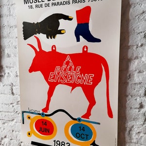 K&ouml;nnte beinhalten: Plakat f&uuml;r das Mus&eacute;e de la Publicit&eacute; mit einer roten Stier-Silhouette und dem Text "A LA BELLE ENSEIGNE". Das Plakat zeigt eine schwarze Hand, einen roten und blauen Stiefel und die Daten: 14. Juni und 14. Oktober 1983.