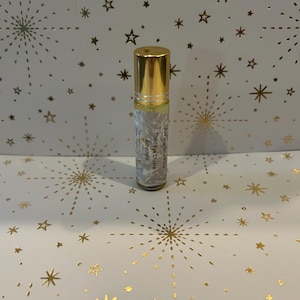 Puede incluir: Un pequeño frasco de vidrio transparente para aceites esenciales con tapa dorada. La etiqueta dice "Third Eye Chakra Amethyst" y "Ava's Scents". El fondo es blanco con un patrón de estrellas doradas.