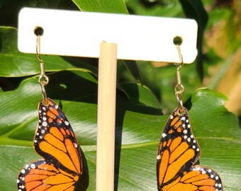May include: A pair of gold butterfly earrings with orange and black wings and white spots. The earrings are displayed on a white card with a wooden dowel.