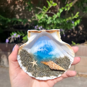 May include: A seashell art piece featuring a miniature beach scene. The shell is filled with sand, a starfish, and a conch shell. A blue and white resin wave design fills the shell, creating a coastal theme.