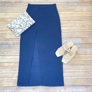 May include: A dark blue maxi skirt with a front slit. The skirt is made of a smooth, stretchy fabric. It is paired with a white clutch and white strappy heels.