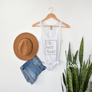 May include: A white tank top with the text "I'd HIIT That" printed on it. The tank top is hanging on a wooden hanger. There is a brown hat, blue denim shorts, and a green plant in the background.
