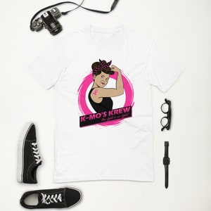 May include: White t-shirt with a pink circle graphic featuring a woman with a pink ribbon tattoo and the text "K-MO'S KREW Her fight is our fight!"