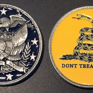 May include: A silver coin with two sides. One side features a bald eagle with an American flag and the words "Liberty or Death". The other side features a yellow snake with the words "Don't Tread on Me".