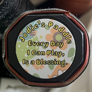 以下が含まれることがあります： 黒い赤いパドルに、白いステッカーが貼ってあり、「Jodie's Paddle Every Day I Can Play, Is a Blessing.」と書かれています。