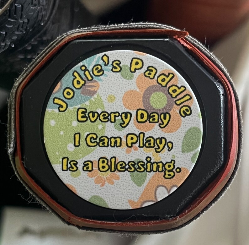 以下が含まれることがあります： 黒い赤いパドルに、白いステッカーが貼ってあり、「Jodie's Paddle Every Day I Can Play, Is a Blessing.」と書かれています。