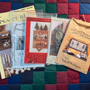 May include: Five different cross-stitch patterns for holiday decor. The patterns include a winter scene with snowmen, a country scene with a wreath, a Christmas scene with carolers, a forest scene with a stocking, and a scene with a bench and geese. The patterns are all from Country Threads.