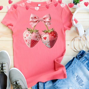 May include: A coral pink t-shirt featuring a strawberry design. The strawberries are made of patchwork fabric with heart and polka dot patterns. A pink bow connects the strawberries. Gray sneakers and blue jeans are also in the image.
