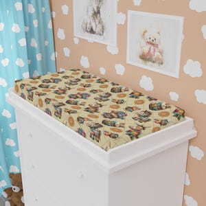 Peut inclure: Une housse de matelas &agrave; langer de couleur cr&egrave;me avec un motif patchwork color&eacute; repr&eacute;sentant des animaux de dessin anim&eacute; et des boutons. La housse est sur une table &agrave; langer blanche avec trois tiroirs.