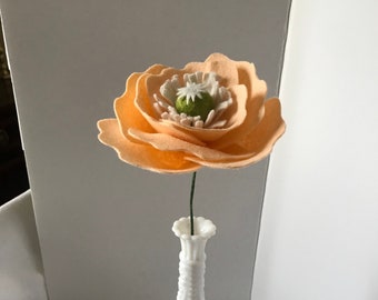 Puede incluir: Una sola flor de fieltro de color melocotón con un centro blanco y un capullo central verde en un jarrón de vidrio de leche blanco.