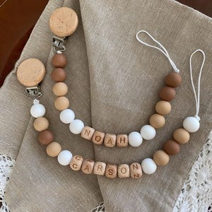 May include: Two baby pacifier clips with wooden beads in shades of brown and white. The clips have wooden disc attachments and spell out the names "Noah" and "Carson" with wooden letter beads. The clips are on a tan cloth.