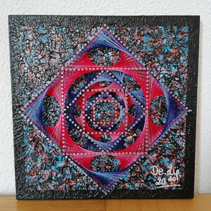 Op de afbeelding: Vierkant string art-stuk met een geometrisch ontwerp in rood, blauw en zilver tegen een getextureerde zwarte achtergrond. Het kunstwerk heeft een centrale spiraal en de handtekening en datum van de kunstenaar.