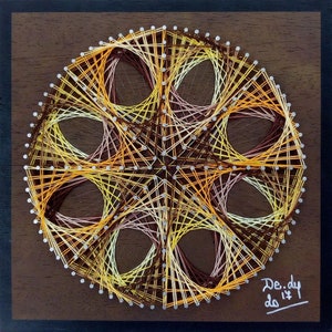 May include: A circular string art piece with a geometric design. The artwork features a pattern of interwoven threads in shades of yellow, orange, and brown, creating a layered effect. The piece is mounted on a dark brown wooden square with a black frame. Signed "De.dy 2017".