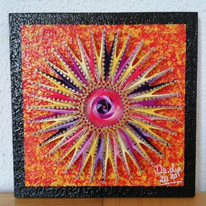 May include: A colorful string art piece featuring a starburst design. The artwork uses pink, yellow, purple, and silver threads against a textured orange background. The piece is framed in black and signed "De.dip 2020".