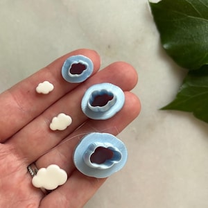 Può includere: Una collezione di taglierini in argilla polimerica a forma di nuvola e pezzi di argilla a forma di nuvola. I taglierini sono azzurri con l'interno rosso scuro. I pezzi di argilla sono bianchi.