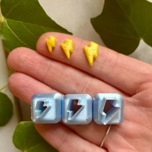 Può includere: Una mano tiene tre piccoli ciondoli a forma di fulmine gialli e tre ciondoli quadrati azzurri con ritagli a forma di fulmine. Sullo sfondo ci sono foglie verdi.