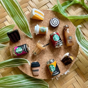 Può includere: Una collezione di miniature di cibi e bevande, tra cui una fetta di torta, una pasticceria al cioccolato, una tazza di caffè con la parola "coffee" e diversi articoli a marchio Starbucks. Gli articoli sono disposti su un sottobicchiere di sughero con foglie verdi e bianche.