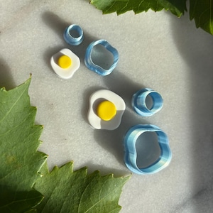 Könnte beinhalten: Eine Sammlung von Polymer Clay Ausstechern in Form von Spiegeleiern und unregelmäßigen Ringen. Die Eierformen haben weiße "Eierweiß" und gelbe "Eigelb". Die Ringausstecher sind hellblau. Grüne Blätter umrahmen die Objekte.