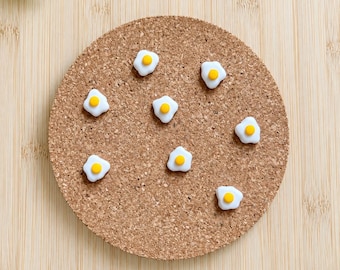 Può includere: Un sottobicchiere rotondo in sughero con sette piccole decorazioni a forma di uovo al tegamino, bianche e gialle, sparse sulla superficie testurizzata color marrone chiaro. Il sottobicchiere ha un design circolare piatto in sughero.