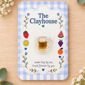 Può includere: Un ciondolo a forma di boccale di birra in resina trasparente in miniatura fissato su un cartoncino rettangolare con bordo a quadretti blu. Il cartoncino riporta il testo The Clayhouse, illustrazioni di frutta e la frase made tiny by me, loved forever by you.