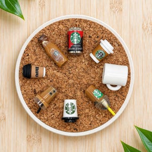 Könnte beinhalten: Eine Sammlung von Miniatur-Kaffeeartikeln, die auf einem runden Korkbrett mit weißem Rand angeordnet sind. Die Gegenstände umfassen eine Starbucks Doubleshot-Dose, verschiedene Kaffeetassen, eine Flasche und einen Becher mit Logos.