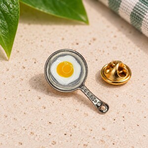 May include: A silver-toned metal lapel pin shaped like a small frying pan containing a fried egg with a bright yellow yolk and white edges. The handle of the pan features a row of small, clear, circular accents. A gold-toned metal clutch back is included.