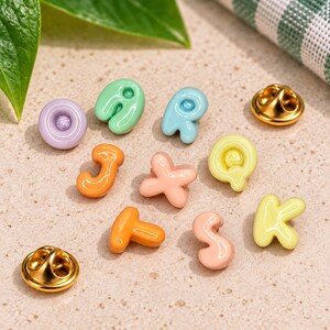 Può includere: Una collezione di piccole spille in smalto lucido dai colori pastello, che raffigurano le lettere O, J, R, Q, T, S, K e X. Ogni spilla presenta una superficie liscia e arrotondata con una chiusura in metallo dorato sul retro.