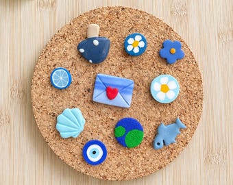 Könnte beinhalten: Eine Sammlung von zehn kleinen, bunten Tonanhängern, die auf einem runden Korkuntersetzer angeordnet sind. Die Anhänger umfassen einen blauen Pilz, eine blaue Blume mit weißer Mitte, einen blauen Umschlag mit rotem Herz, eine Zitrusfrucht, eine Muschel und einen Fisch.
