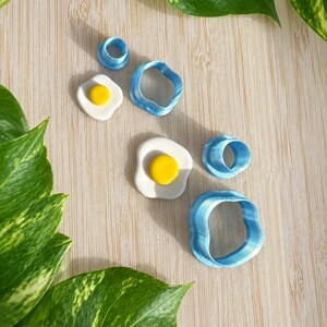 May include: Two sets of blue plastic cutters and two corresponding fried egg shapes made of white and yellow material. Each set includes a circular cutter for the yolk and an irregular shaped cutter for the egg white, creating a sunny side up egg.