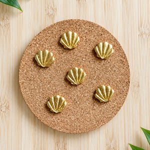 May include: Six small, gold-coloured metal seashell-shaped pins or decorative accents arranged in a circular pattern on a round, textured, light brown cork board. Each piece features a fan-like design with distinct ridges radiating from the base.