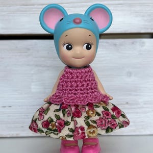 Puede incluir: Una peque&ntilde;a mu&ntilde;eca con un gorro de rat&oacute;n azul con orejas rosas, una parte superior de ganchillo rosa y una falda con estampado floral. La mu&ntilde;eca lleva zapatos rosas y tiene una expresi&oacute;n sonriente.
