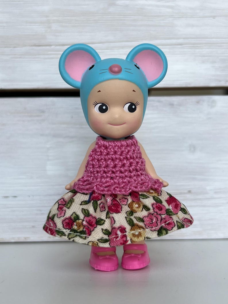 Puede incluir: Una peque&ntilde;a mu&ntilde;eca con un gorro de rat&oacute;n azul con orejas rosas, una parte superior de ganchillo rosa y una falda con estampado floral. La mu&ntilde;eca lleva zapatos rosas y tiene una expresi&oacute;n sonriente.