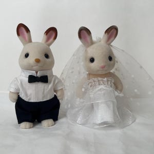 May include: Two flocked rabbit figurines dressed for a wedding. The left figure wears a white collared shirt with a black bow tie and dark trousers. The right figure wears a white dress with a layered skirt and a sheer veil with white polka dots.