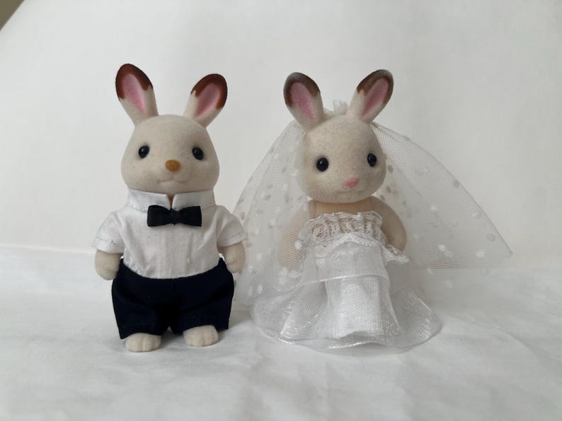 May include: Two flocked rabbit figurines dressed for a wedding. The left figure wears a white collared shirt with a black bow tie and dark trousers. The right figure wears a white dress with a layered skirt and a sheer veil with white polka dots.