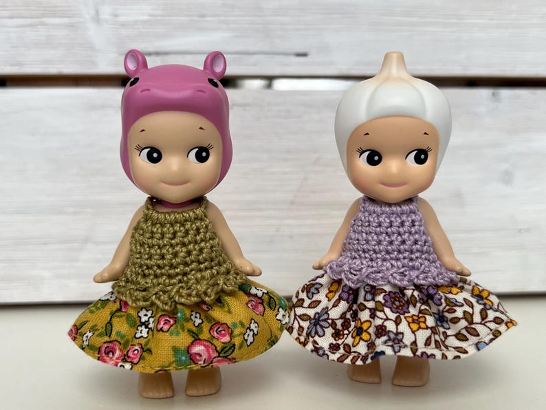 Puede incluir: Dos peque&ntilde;as figuras, una con cabeza de hipop&oacute;tamo rosa y otra con cabeza de bulbo de ajo. Ambas visten tops de ganchillo y faldas con estampado floral. La falda de la figura del hipop&oacute;tamo es amarilla con rosas rosas, mientras que la falda de la figura del ajo es blanca con flores moradas y amarillas.