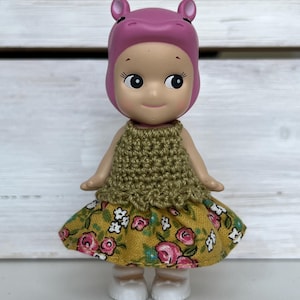 Puede incluir: Una peque&ntilde;a figura de un personaje con tem&aacute;tica de hipop&oacute;tamo que lleva una capucha rosa, una parte superior de ganchillo verde oliva y una falda amarilla con estampados florales rosas. La figura tiene zapatos blancos y una expresi&oacute;n sonriente.