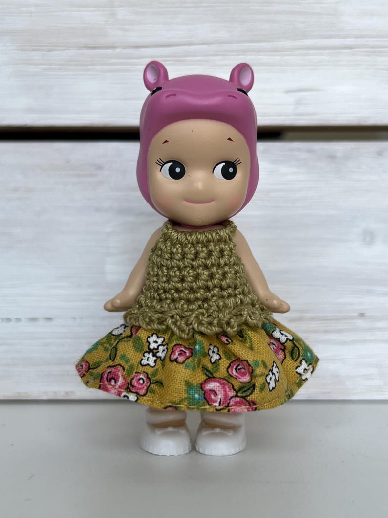 Puede incluir: Una peque&ntilde;a figura de un personaje con tem&aacute;tica de hipop&oacute;tamo que lleva una capucha rosa, una parte superior de ganchillo verde oliva y una falda amarilla con estampados florales rosas. La figura tiene zapatos blancos y una expresi&oacute;n sonriente.