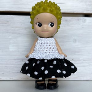 Puede incluir: Una peque&ntilde;a mu&ntilde;eca con una cabeza amarilla puntiaguda, que lleva un top blanco de ganchillo y una falda negra con lunares blancos. La mu&ntilde;eca tiene zapatos negros y est&aacute; de pie frente a un fondo de madera blanca.