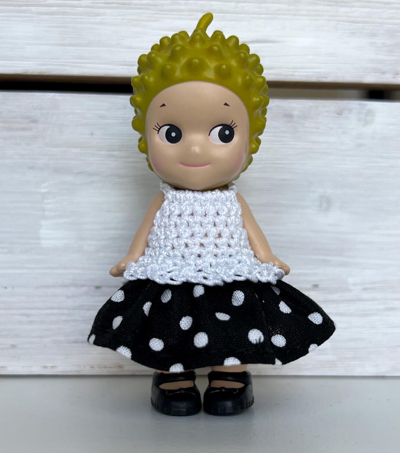 Puede incluir: Una peque&ntilde;a mu&ntilde;eca con una cabeza amarilla puntiaguda, que lleva un top blanco de ganchillo y una falda negra con lunares blancos. La mu&ntilde;eca tiene zapatos negros y est&aacute; de pie frente a un fondo de madera blanca.