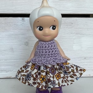 Puede incluir: Una mu&ntilde;eca caprichosa con una cabeza blanca bulbosa y una cara sonriente. Viste un top de ganchillo lavanda y una falda con estampado floral, con zapatos morados. Un objeto de colecci&oacute;n encantador.