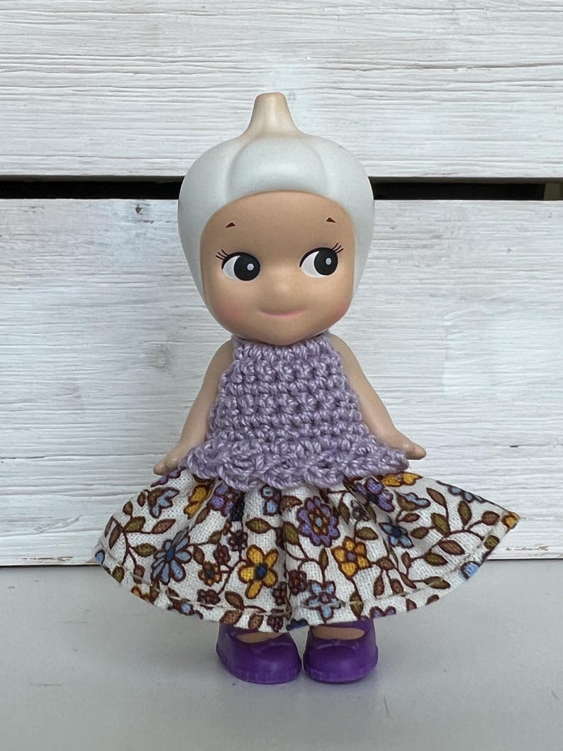 Puede incluir: Una mu&ntilde;eca caprichosa con una cabeza blanca bulbosa y una cara sonriente. Viste un top de ganchillo lavanda y una falda con estampado floral, con zapatos morados. Un objeto de colecci&oacute;n encantador.